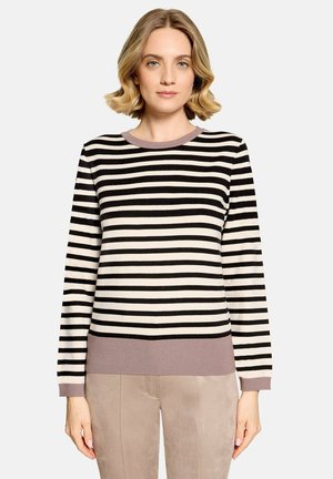 Kvinde med kort blondt hår iført en sort og hvid stribet sweater med mauve kant og lyse beige bukser, stående mod en ensfarvet baggrund.