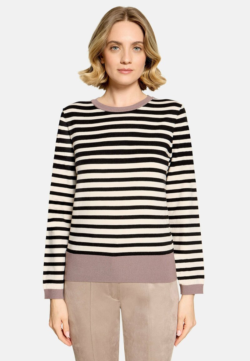 Femme aux cheveux blonds courts portant un pull rayé noir et blanc avec une bordure mauve et un pantalon beige clair, debout devant un fond uni.