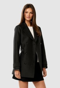 Woman with long brown hair wearing a black double-breasted blazer over a white top and black skirt, standing against a plain background.