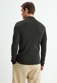 Dunkelgrauer gerippter Pullover mit hohem Kragen und taillierter Passform, hat lange Ärmel und einen geraden Saum, getragen mit hellbeigen Hosen.