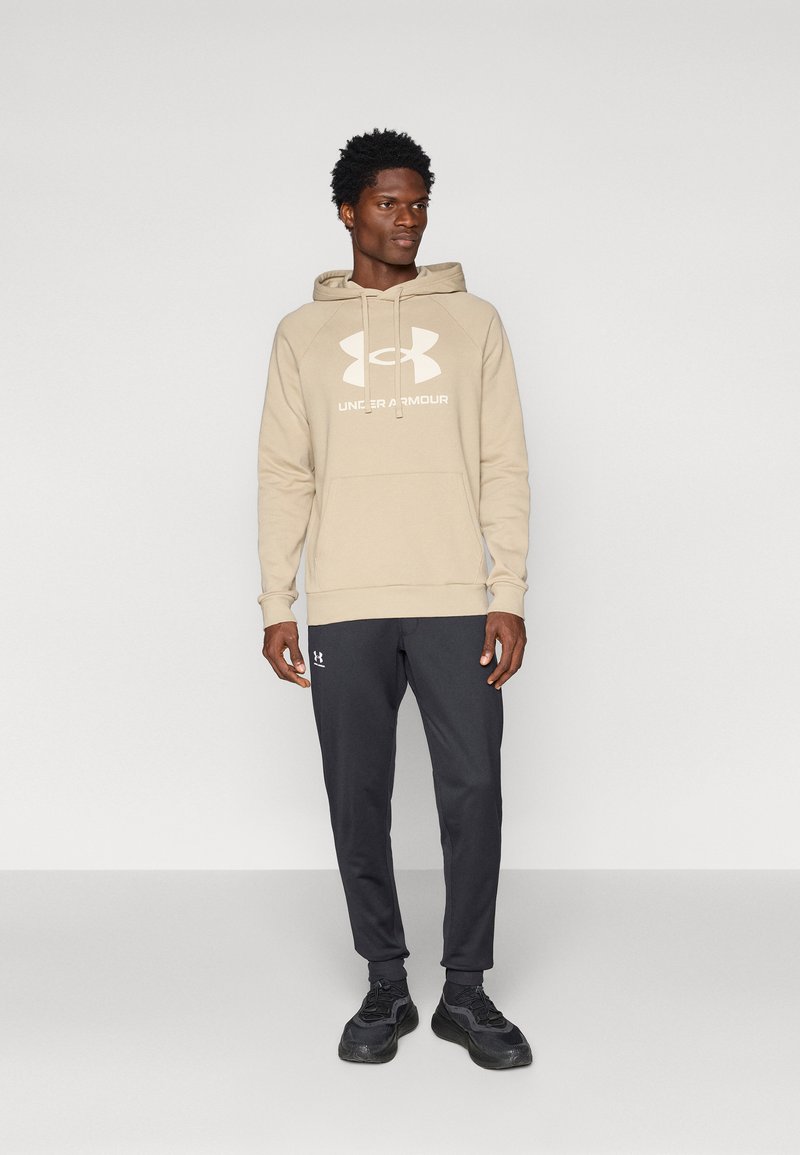 Felpa beige con un grande logo bianco, abbinata a pantaloni sportivi neri. La felpa ha una tasca a kangaroo e un cappuccio con cordino.
