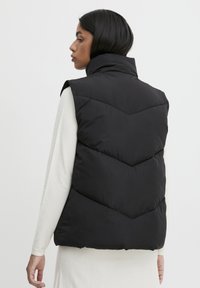 Gilet matelassé noir avec un col montant, présentant un motif matelassé en chevrons. Le modèle porte une chemise à manches longues crème en dessous.