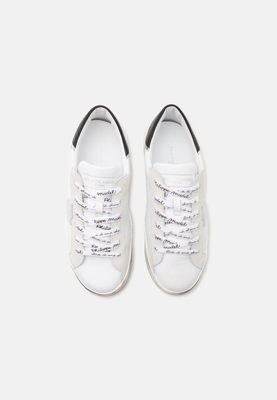 Paire de baskets blanches avec détails noirs au talon et lacets blancs imprimés de texte noir en écriture cursive, sur un fond uni clair.