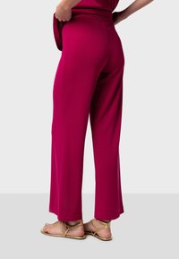 Pantalons larges fuchsia en tissu lisse, dotés d'une taille haute. Associés à des sandales métalliques dorées, montrant une vue arrière.