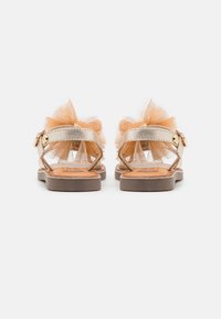 Złote płaskie sandały z paskami na kostkę i dużymi brązowymi kokardami z tiulu z tyłu, pokazane od tyłu na białym tle.