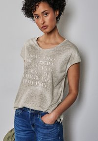 Camiseta beige con un patrón sutil, cuello redondo y mangas cortas. En la parte frontal presenta texto blanco con un acabado brillante. Combinada con jeans azules.