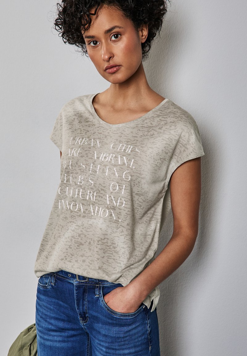 Camiseta beige con un patrón sutil, cuello redondo y mangas cortas. En la parte frontal presenta texto blanco con un acabado brillante. Combinada con jeans azules.