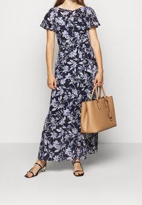 Kvinna klädd i en marinblå blommig maxiklänning med svävande ärmar, svarta sandaler med remmar och bärande en stor handväska i tanläder.