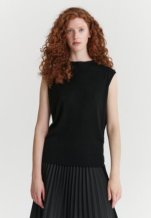 Femme aux longs cheveux roux bouclés portant un haut noir sans manches et une jupe plissée noire, debout devant un fond blanc uni.