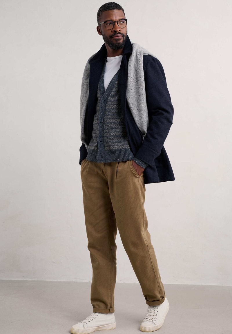 Herenoutfit met een marineblauwe jas met grijze schouderaccenten, een grijs patrooncardigan, een witte t-shirt, bruine broek en witte sneakers.