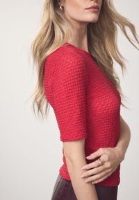 Crveni teksturirani top s kratkim rukavima i uskim krojem. Tkanina ima istaknut uzorak i kombinirana je s tamnim kožnim hlačama.