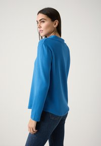 Langsleeve pullover in helder blauw, met een eenvoudige kraag, zachte textuur en een relaxte pasvorm. Draagt met donkerdenim jeans.