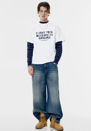 Tricou alb din bumbac cu text negru, suprapus peste o cămașă cu mânecă lungă în dungi bleumarin, asociat cu blugi lejeri din denim albastru și cizme galbene.