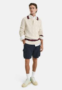 Creme kabelstrik sweater med en kontrastfarvet navy og rød stribet krave, parret med navy cargo shorts og hvide sneakers med grønne detaljer.