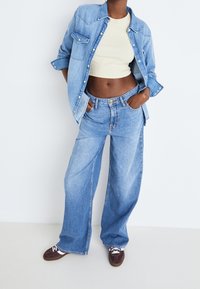 Světle modrá džínová bunda s detaily knoflíků, v kombinaci se světle modrými kalhotami s vysokým pasem a širokými nohavicemi, nošena s tmavě hnědými botami.