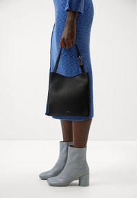 Sac fourre-tout en cuir texturé noir avec un sommet incurvé, tenu par une main. Porté avec une robe bleu à côtes et des bottines bleu clair avec un talon carré.