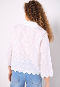 Blusa blanca bordada con un patrón floral, dobladillo en forma de escalloped y mangas de tres cuartos, llevada con unos jeans de mezclilla azules.