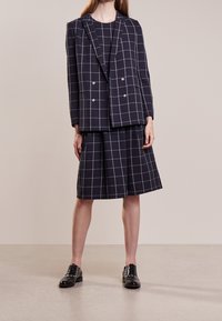 Marinblå rutigt kostym består av en dubbelknäppt blazer och en plisserad kjol, tillverkad av texturerad tyg, i kombination med svarta snörskor.