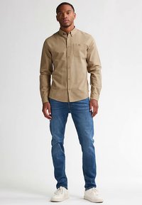 Beżowa koszula na guziki z kieszenią na piersi, zestawiona z niebieskimi dżinsami i białymi trampkami. Koszula ma klasyczny kołnierzyk i standardowy krój.