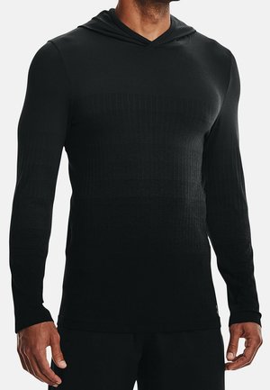 Hombre vistiendo una camiseta negra de manga larga y capucha ajustada con un sutil patrón texturizado, de pie frente a un fondo claro y liso.