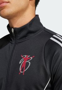 Chaqueta negra con cremallera y cuello alto, detalles en gris y un logotipo gráfico rojo en el pecho. Presenta rayas blancas en los hombros.