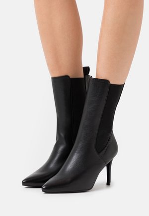 Black leather mid-calf boots with pointed toes, elastic side panels, and slim high heels worn on bare legs against a white background.