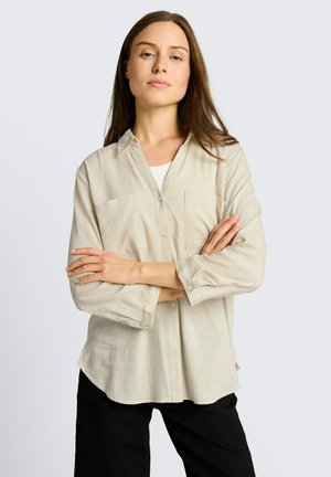 Kobieta z długimi brązowymi włosami, ubrana w beżową koszulę z guzikami na białą bluzkę, czarne spodnie, stoi ze skrzyżowanymi ramionami na jednolitym jasnym tle.