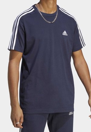Personne portant un t-shirt Adidas bleu marine à manches courtes avec des détails à trois bandes blanches sur les épaules et le logo Adidas sur la poitrine.