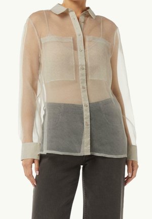 Femme portant un chemisier beige transparent à manches longues boutonné avec des poches visibles sur un top, associé à un pantalon gris foncé.