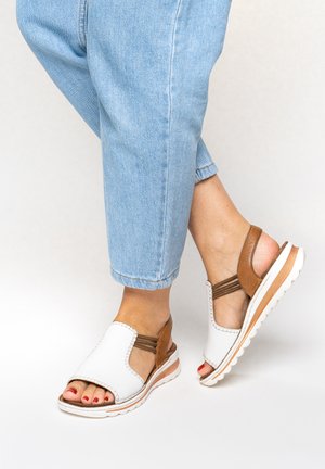 Pieds chaussés de sandales ouvertes blanches et beige avec des sangles élastiques, associés à un jean bleu clair coupé court sur un fond blanc.