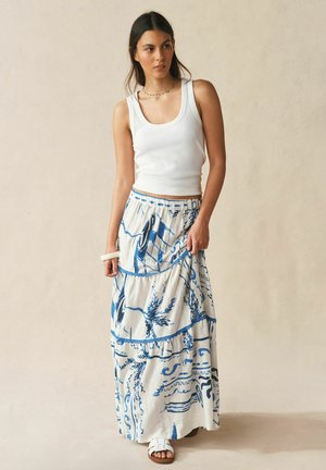 Fehér ujjatlan felsőt, kék botanikus mintás, hosszú fehér maxiszoknyát és fehér papucsot viselő nő bézs háttér előtt áll.