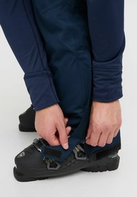 Pantalones de esquí azul marino con una textura suave, costuras detalladas y cierre ajustable cerca de botas de esquí negras con herrajes metálicos.
