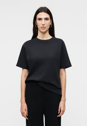 Γυναίκα με μακριά σκούρα μαλλιά, που φοράει χρυσούς κρίκους στα αυτιά, μαύρη κοντομάνικη μπλούζα και μαύρο παντελόνι, στέκεται μπροστά σε απλό λευκό φόντο.