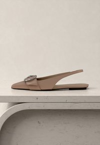 Μπεζ πλατφόρμα τύπου slingback με μυτερή μύτη και μεγάλο ασημένιο πόρπη, τοποθετημένη σε ανοιχτή μαρμάρινη επιφάνεια με ουδέτερο φόντο.