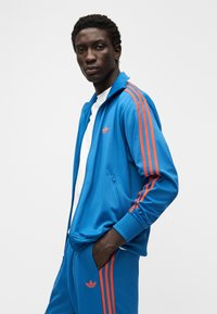 Μπλε αθλητική φόρμα με κόκκινες τρεις ρίγες, ζακέτα με φερμουάρ και παντελόνι που ταιριάζει. Το υλικό φαίνεται λείο, με λογότυπο Adidas.