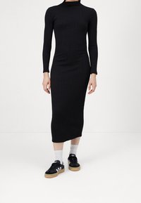 Abito midi nero a costine con maniche lunghe e collo alto, abbinato a sneaker nere con strisce bianche e suola in gomma.