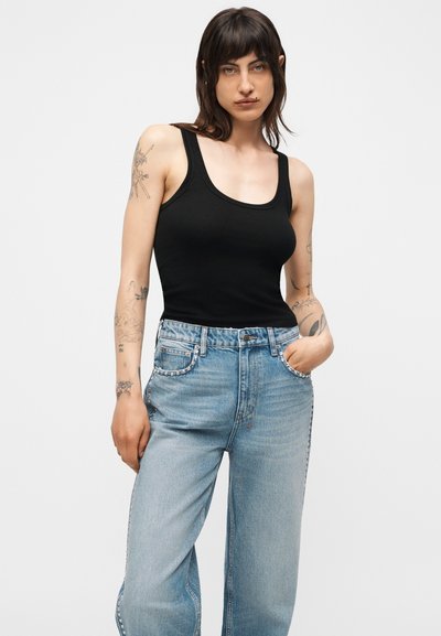 Γυναίκα με σκούρα μαλλιά και τατουάζ που φοράει ένα μαύρο μπλουζάκι χωρίς μανίκια και ανοιχτό μπλε τζιν με ψηλή μέση και διακοσμητικά με στρας.