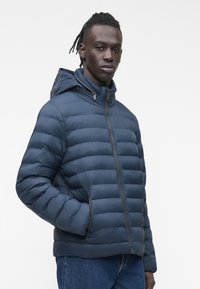 Vīrietis ar tumšu ādu un bizētiem matiem, valkājot tumši zilu kapuci ar vates odējumu un zilas džinsas, stāv ar vienu roku kabatā.