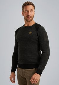 Man in een donkergrijze gebreide trui met schouderpatches en een klein borstellogo, gecombineerd met een paar bruine broek, die naar de zijkant kijkt.