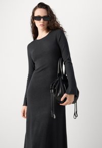 Long-sleeve, fitted charcoal gray dress with ribbed texture. Model holds a black, cinched handbag with drawstring details. Dark sunglasses worn.