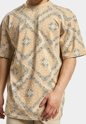 Mężczyzna w beżowej koszuli z krótkim rękawem z kremowo-czarnym wzorem paisley i kwiatowym na bandanie, dłoń spoczywa blisko talii.