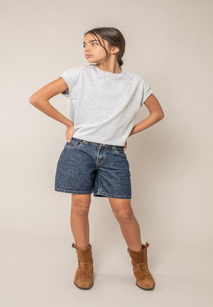 Jonge vrouw staand met handen in de zij, gekleed in een grijs T-shirt, denim korte broek en bruine suède enkellaarzen tegen een effen achtergrond.