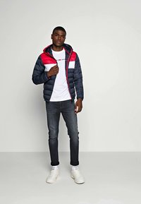 Marineblauwe en rode donsjack met een ritssluiting aan de voorkant, witte paneel en elastische manchetten, gedragen over een wit T-shirt en donkere jeans, gecombineerd met witte sneakers.