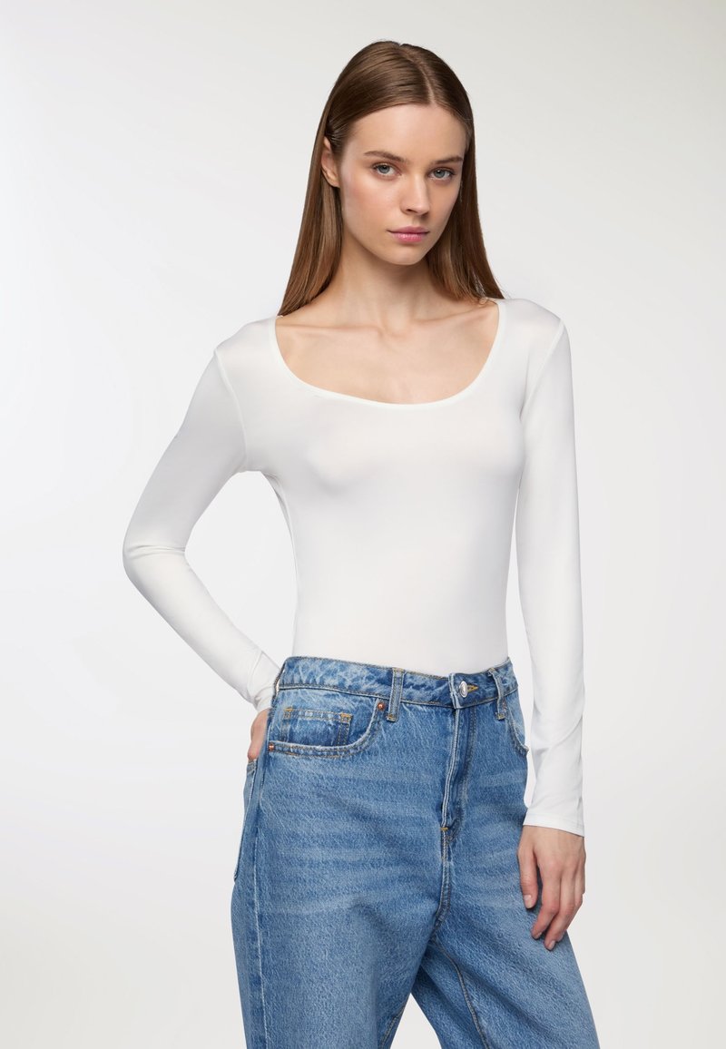 Biały bodysuit z długim rękawem i szerokim dekoltem w serek, zestawiony z niebieskimi dżinsami o wysokim stanie. Gładka tekstura i dopasowany krój.