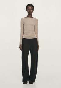 Donna in piedi che indossa una maglietta fitted a maniche lunghe beige e pantaloni neri a gamba larga davanti a uno sfondo chiaro e neutro.