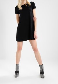 Černé šaty s krátkým rukávem a volným střihem, vyrobené z jemné látky. Nosené s stříbrnými třpytivými kotníkovými botami s černými popruhy.