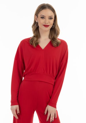Rød jumpsuit med V-udskæring, lange ærmer og samlet talje. Glat tekstur med et tætsiddende snit og enkle, rene linjer.