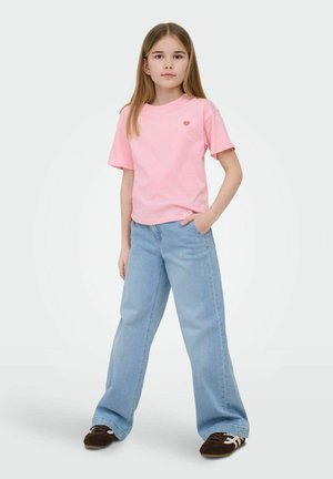 Jeune fille debout, une main dans la poche, portant un t-shirt rose clair avec un petit cœur et un jean large bleu clair.