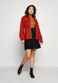 Sieviete, apģērbta rūsu krāsas zipa jaku virs brūna turtlenecka, melnas pludinātas svārkus un tumšas potīšu zābakus, stāv pret vienkāršu fonu.