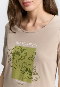 Beige T-Shirt mit einem grünen Blumenmuster, rundem Halsausschnitt und kurzen Ärmeln. Der Text enthält "BLOOMING" und "flowers forever" in Weiß.
